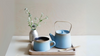 Blue ceramic teaware set, featuring a small coffee mug and a matching blue teapot. The teaware sits on a small wooden tray, staged with a small bamboo spoon and a flower arrangement in a silver vase.